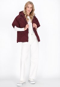 Vesta puffer matlasată de culoare burgundă, cu glugă, mâneci scurte și închidere cu fermoar. Purtată peste o cămașă albă cu mâneci lungi și combinată cu pantaloni albi.