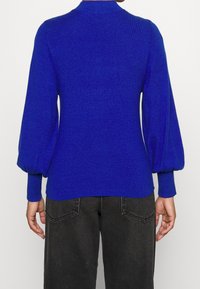 Maglione blu lavorato a maglia con collo alto, maniche a palloncino e polsini a costine. Indossato con pantaloni scuri a vita alta, che mostrano una texture liscia.