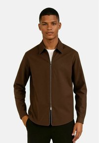 Brun zip-up jakke med krage, lange ermer og en jevn tekstur. Har en sølvglidelås og subtil logo detalj på brystet.