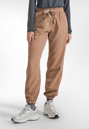 Persoon die lichtbruine broek met trekkoord, grijze sokken, zilveren sneakers en een donkergrijs shirt met lange mouwen draagt, staand op een witte achtergrond.