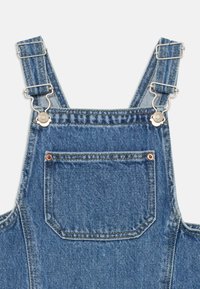 Denim kopējais ar regulējamu sudraba aparatūru, priekšējo kabatu un klasisku zilo mazgāšanu ar subtīliem izbalēšanas un šuvju rakstiem.