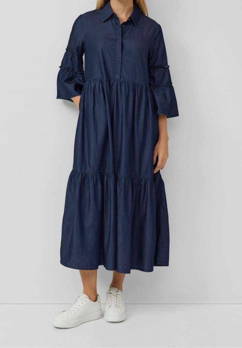 Femme portant une longue robe bleu marine à jupe à étages et manches à volants longueur coude, associée à des baskets blanches.