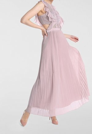 Frau trägt ein ärmelloses, blassrosa Kleid mit Volants, Häkeloberteil, seitlichen Ausschnitten und plissiertem Rock, kombiniert mit passenden High Heels.