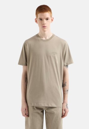 Camiseta de algodón beige con cuello redondo, mangas cortas y un pequeño logo estampado en el pecho. Presenta un corte relajado y una textura suave.