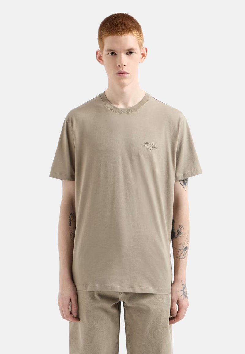 Beiges Baumwoll-T-Shirt mit Rundhalsausschnitt, kurzen Ärmeln und kleinem aufgedrucktem Logo auf der Brust. Es hat eine lockere Passform und eine glatte Textur.