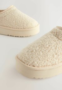 Zapatos de slip-on color crema con parte superior texturizada y suave, y suela de goma de color claro. Presentan un perfil bajo y puntera redondeada.