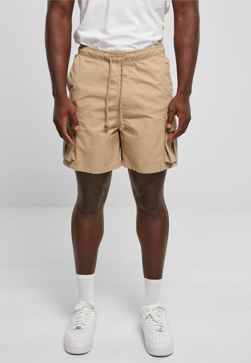 Man wearing beige cargo shorts, white socks, and white sneakers standing against a plain gray background.