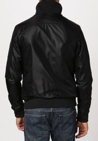 Svart bomberjacka i ett mjukt och lätt tyg, med hög krage och ribbade manschetter. Visad bakifrån, med dragkedjedetaljer synliga.