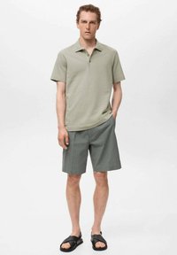 Ljusgrön pikétröja med campkrage, kombinerad med grå rutiga shorts. Modellen har svarta sandaler. Slät bomullstextur.