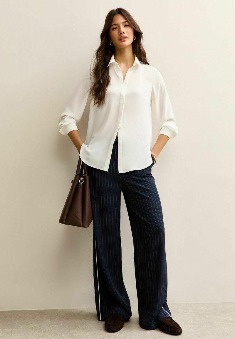 Blusa blanca de manga larga con botones, combinada con pantalones anchos de rayas finas en azul marino. También se incluye un bolso marrón.