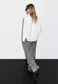 Camicia bianca oversize abbinata a pantaloni grigi a gamba larga e tacchi scuri. La camicia ha polsini con bottoni e il tessuto appare liscio.