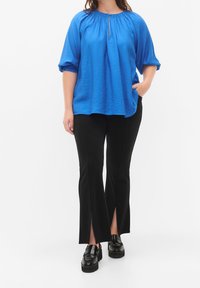 Blusa azul texturizada con detalle de escote en forma de llave, combinada con pantalones negros con aberturas laterales. El modelo está de pie con una mano en el bolsillo.