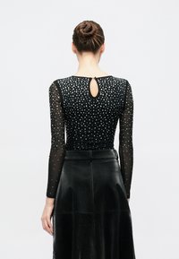 Μαύρο κορμάκι με μακριά διαφανή μανίκια, διακοσμημένο με ασημένιες τρουκς. Διαθέτει λεπτομέρεια πίσω με άνοιγμα σε σχήμα κλειδιού. Συνδυασμένο με μαύρη δερμάτινη φούστα.