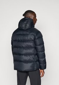 Doudoune bleu marine avec une capuche, présentant un design large, une texture matelassée et une finition lisse et brillante. La vue arrière met en avant son volume.