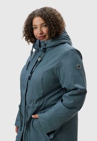 Modrá vodotěsná parka s kapucí, nastavitelné šnůrky, boční kapsy a kulatý detail na rukávu. Hladký, lesklý materiál.