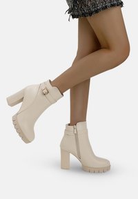 Botines beige con tacón grueso, material sintético suave, cremallera lateral y detalle de tira con hebilla cuadrada. Suela de goma gruesa.