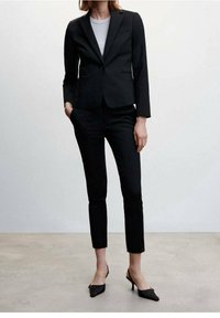 Zwarte op maat gemaakte blazer met een enkele knop, gecombineerd met strakke zwarte broeken. De outfit wordt aangevuld met puntige zwarte hakken.