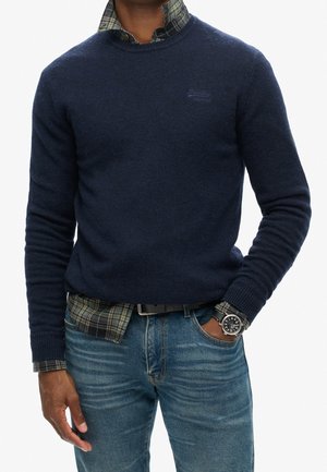 Mann trägt einen marineblauen Pullover über einem grünen karierten Hemd, blaue Jeans, schwarzen Gürtel und eine silberne Armbanduhr mit schwarzem Zifferblatt, Hand in der Tasche.