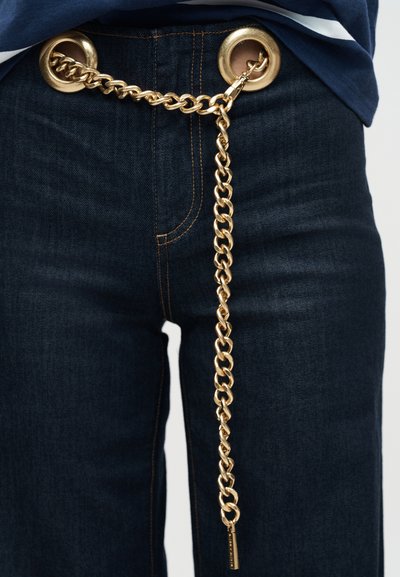 Χρυσή αλυσίδα ζώνης περασμένη μέσα από μεγάλα χρυσά κουμπώματα σε σκούρο μπλε τζιν, φορεμένη με μπλε τοπ.