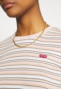 Pruhované tričko s horizontálními pruhy v růžové, bílé a černé barvě. Obsahuje malé červené logo Levi's na levé hrudi a zlatý řetízek.