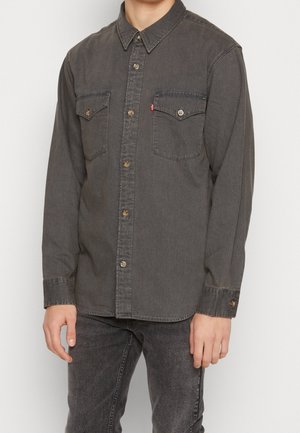 Mies pukeutuneena haalistuneeseen mustaan farkkupaitaan, jossa on kaksi rintataskua, ja mustiin farkkuihin, seisoo kädet rennosti sivuilla.