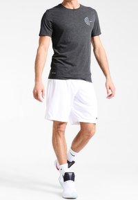 Mężczyzna ubrany w ciemnoszary t-shirt Nike, białe spodenki Nike, białe skarpetki Nike i białe sneakersy z czerwonymi i czarnymi detalami, stojący na białym tle.