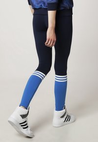 Námorně modré legíny s třemi bílými pruhy na lýtkách, vyrobené z elastické tkaniny, spárované s bílými teniskami s černými akcenty.