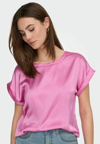 Růžový saténový top s krátkými rukávy s ohrnutím, kulatým výstřihem a volným střihem, kombinovaný se světle modrými džínami.