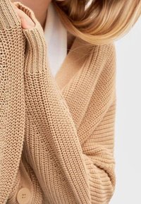 Jasnobrązowy sweter z dzianiny o prążkowanej fakturze i długich rękawach, z zapięciem na guziki i głębokim dekoltem w kształcie litery V.