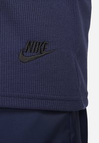 Tmavě modré texturované tričko s krátkým rukávem, které má na levé straně černě vyšívané logo Nike, s rovním lemem a kulatým výstřihem.