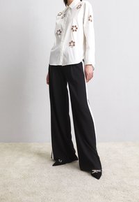 Vit skjorta med blommig broderi; parat med svarta vida byxor med vita sidostripes och spetsiga svarta skor.