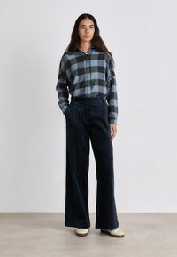 Plaid shirt in blue and black hues, with a buttoned collar. Paired with dark, wide-leg corduroy pants and light-colored shoes.