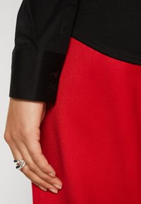 Polsino di camicia nera a maniche lunghe con dettaglio dei bottoni, abbinato a una gonna in tessuto rosso texturizzato. Mano posata sulla gonna, con un anello d'argento.