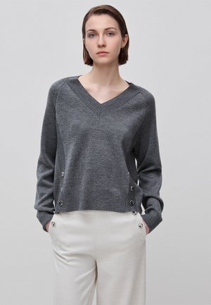 Donna con capelli castani corti che indossa un maglione grigio con scollo a V e bottoni laterali e pantaloni bianchi, in piedi davanti a uno sfondo chiaro e semplice.