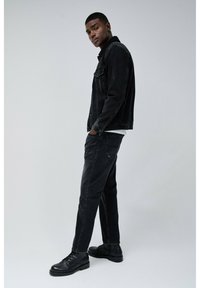 Chaqueta de mezclilla negra con cuello de sherpa, combinada con pantalones negros a juego y botas negras de cordones, sobre un fondo neutro.