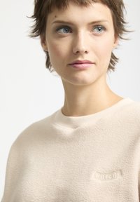 Beżowy polarowy sweatshirt z prążkowanym dekoltem, z haftowanym logo na piersi i miękką, teksturowaną powierzchnią.