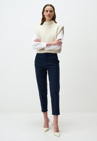 Beige geribbelde mouwloze trui over een witte blouse, gecombineerd met marineblauwe cropped broek en crèmekleurige hoge hakken. Eenvoudig ontwerp, effen kleuren.