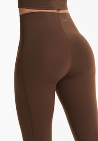 Barna, magas derekú leggings rugalmas anyagból. Simább textúra, testhezálló kialakítás, finom varrási részletek. Márkájának logója látható.