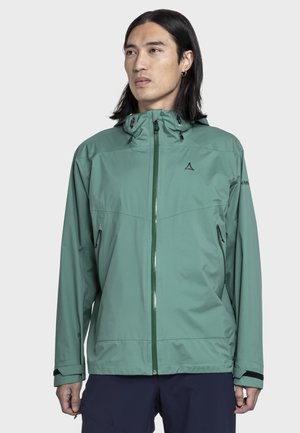 Mann mit langen dunklen Haaren trägt grüne wasserdichte Kapuzenjacke mit Reißverschlusstaschen und schwarze Hose, schaut leicht zur Seite.