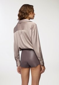 Camicia in satin beige chiaro con retro più scuro a contrasto, abbinata a shorts aderenti marrone scuro, che mostrano texture lisce e un design sartoriale.