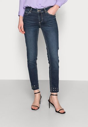 Dark blue skinny jeans with stud accents at the hems, featuring a classic five-pocket design and contrast stitching. Pair with black sandals.