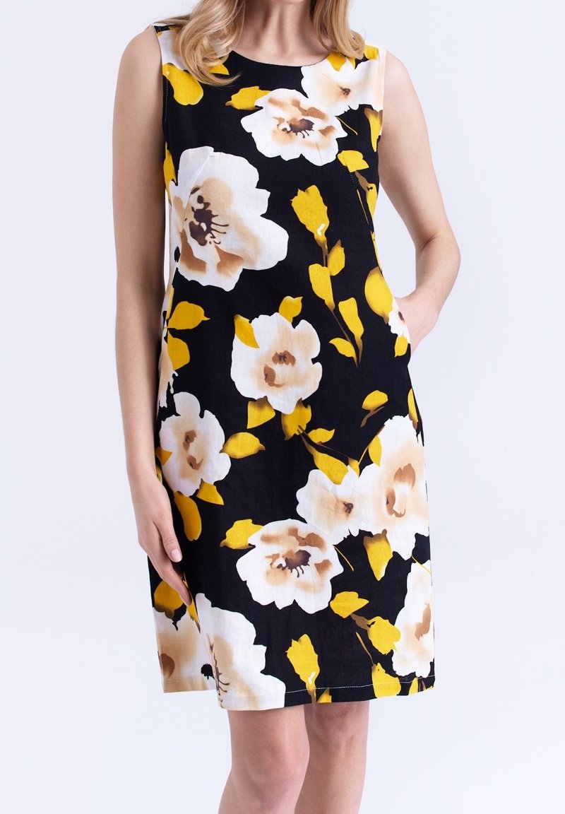 Femme portant une robe noire sans manches avec un grand motif floral blanc et jaune, debout, une main dans la poche.