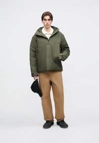 Alyvuogių žalumos striukė su kapišonu, užtrauktuku užsegama, dvi rankų kišenės. Rudos plačios kelnės, juoda kepuraitė rankoje ir juodos avalynės. Paprastas dizainas.