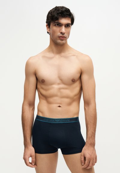 Mannequin masculin aux cheveux courts et foncés portant un boxer noir Emporio Armani, debout devant un fond clair uni.
