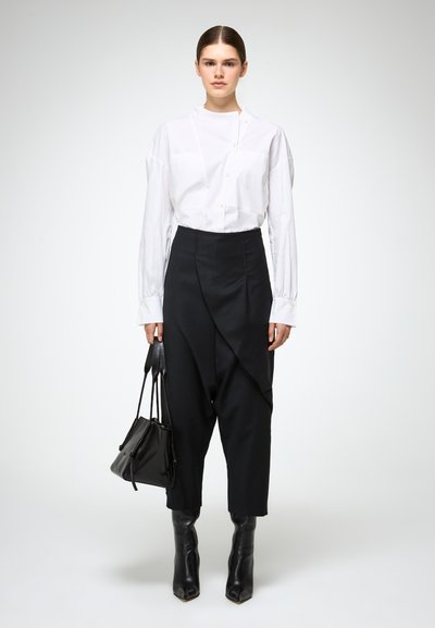 Witte blouse met hoge kraag, gecombineerd met op maat gemaakte zwarte broek met gedrapeerde details en ingeklapte broekspijpen tot de ankle, aangevuld met zwarte laarzen en tas.