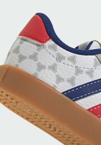 Primer plano del talón de una zapatilla blanca con patrón geométrico gris, pestaña del talón roja, ribete azul marino, rayas azules y rojas, y suela de goma color caramelo.