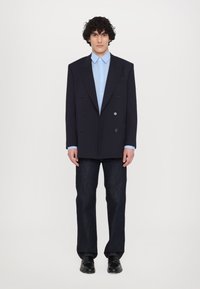 Veste bleu marine croisée avec quatre boutons sur le devant, associée à une chemise bleu clair et un jean en denim foncé, complétée par des chaussures noires.