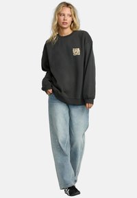 Felpa oversized in carbone con un piccolo logo beige sul petto, abbinata a jeans larghi azzurri e sneakers nere.