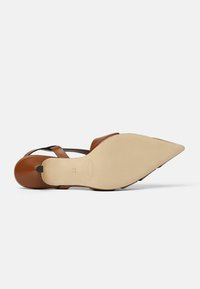 Sandal in pelle marrone con cinturini incrociati, punta appuntita e suola beige liscia con texture antiscivolo e dettaglio del marchio in rilievo.