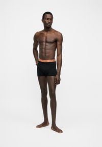 Λεπτός άνδρας με σκουρόχρωμο δέρμα, στέκεται ξυπόλυτος, φοράει μαύρα μπόξερ με πορτοκαλί ζώνη μέσης, σε απλό λευκό φόντο.